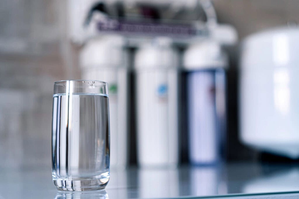 A water glass sits on a counter with a whole-home water treatment system in the background.